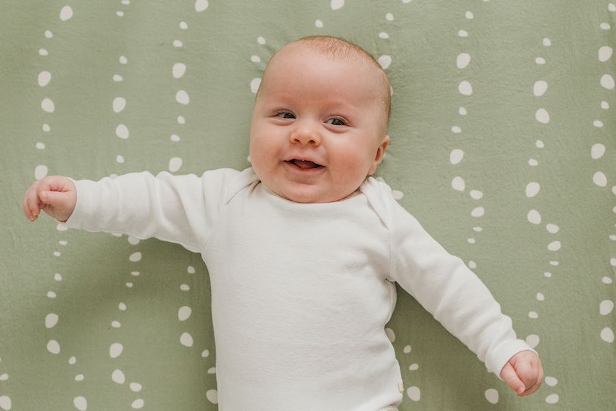 A smiling baby waving his arms, lying down on an Oolie organic cotton crib sheet, printed with a green pattern of white stones or bubbles.