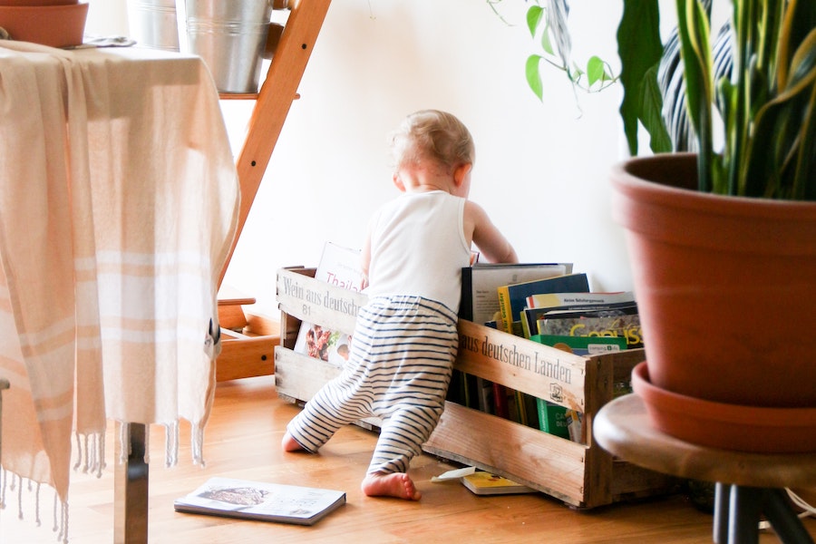 Toddler leaning over a crate filled with books and magazines, surrounded by green houseplants