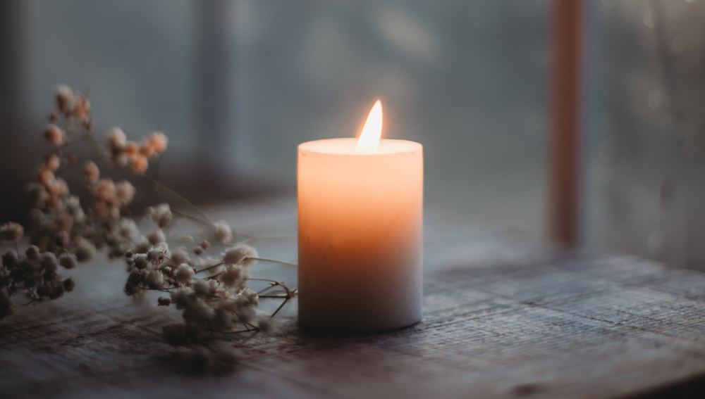 A lit candle glowing warmly on a wooden table top next to some dried flowers