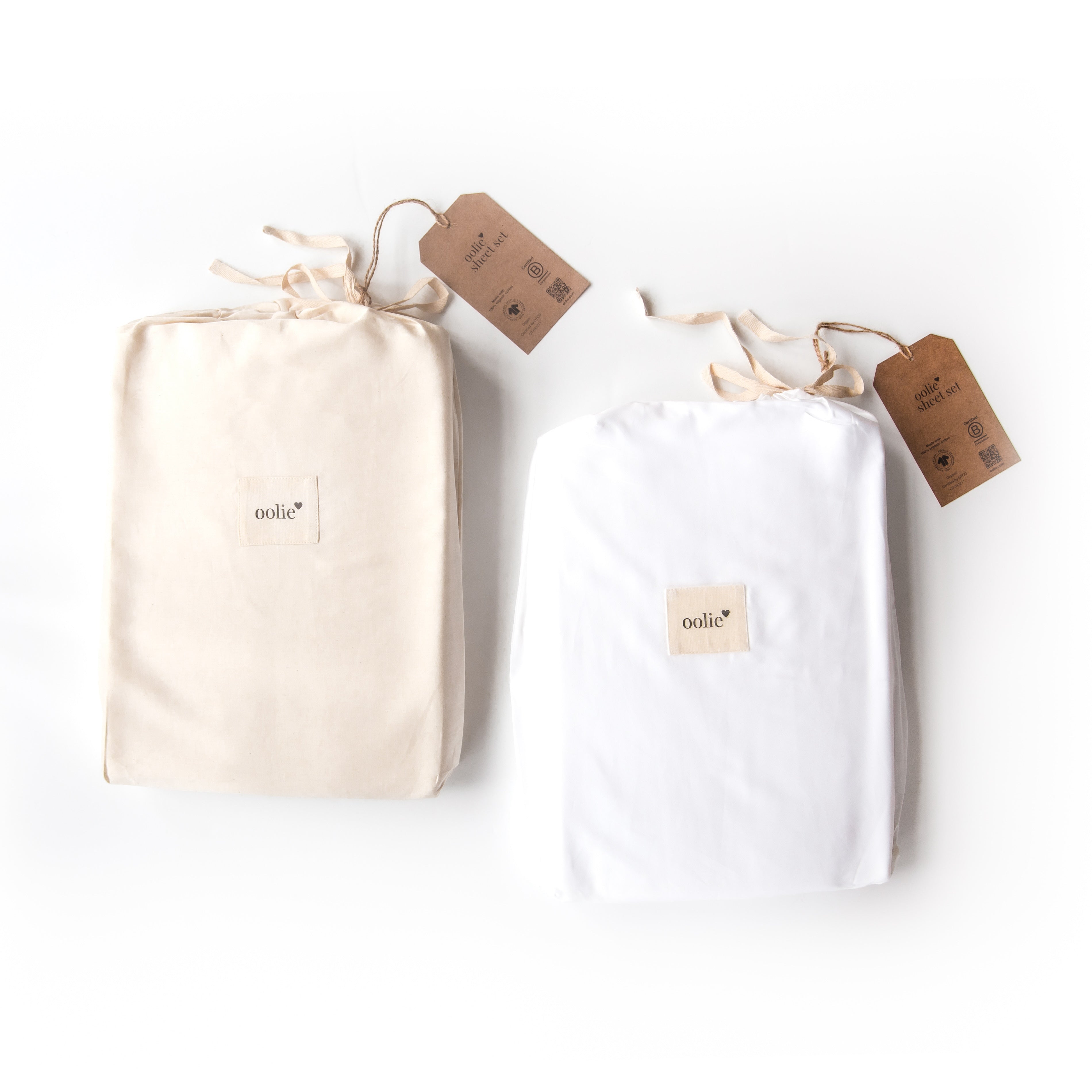 Two Oolie organic cotton sheet sets, one natural and one white, shown on a white background. Both are folded and packaged in fabric bags with recycled kraft paper hang tags.