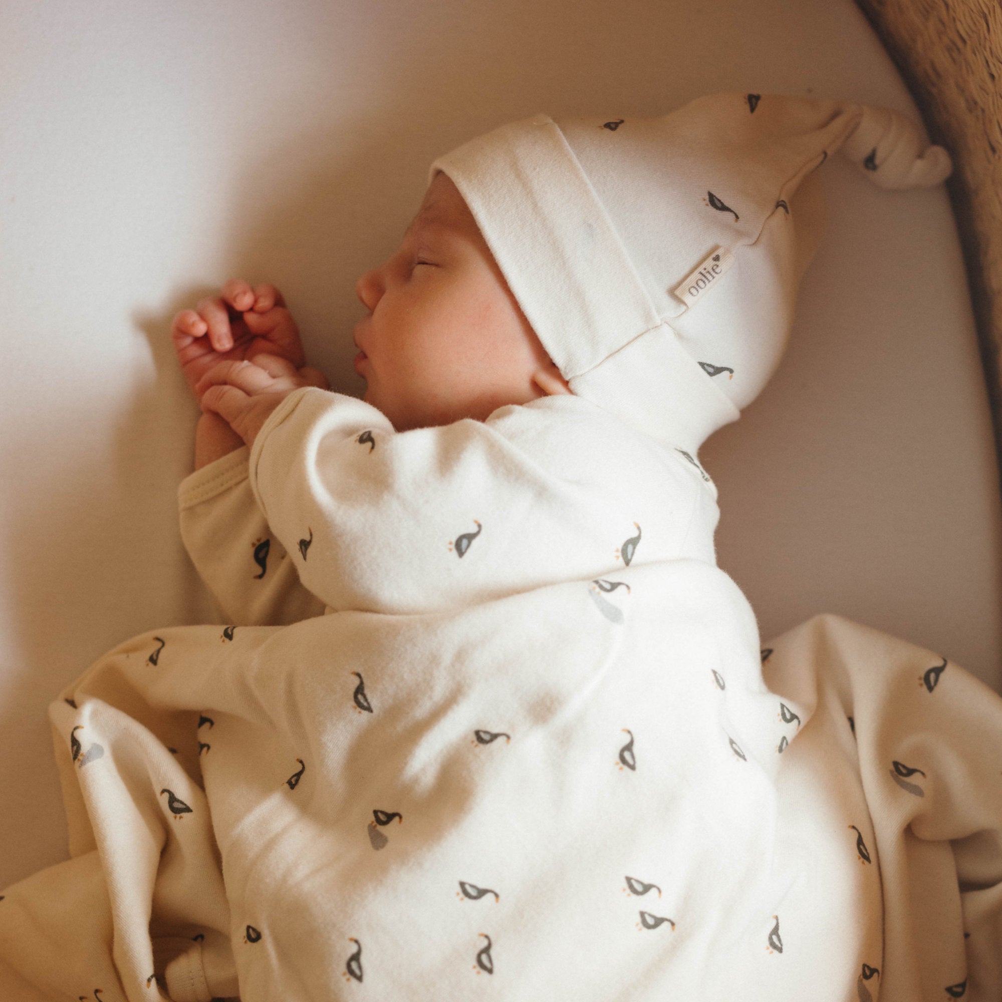 A newborn sleeping gently while wearing an organic cotton hat and bodysuit, draped with an Oolie baby blanket. The hat, bodysuit, and blanket all have a repeating printed pattern of small ducks.