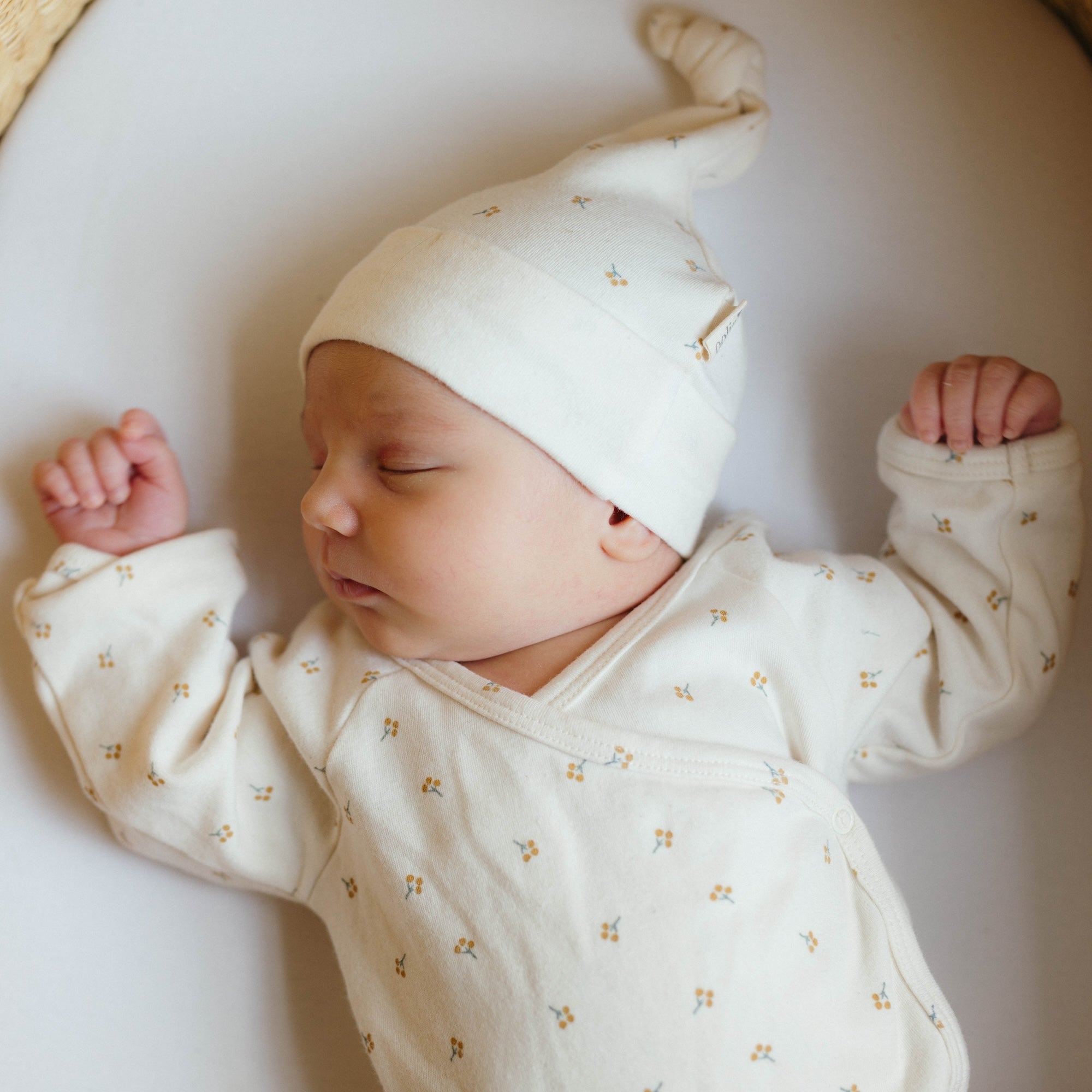 A newborn baby sleeping in a bassinet. The baby is wearing an organic cotton baby hat and an organic cotton bodysuit from Oolie. Both the blanket and the hat have a repeating printed pattern of a small sprig with golden berries.