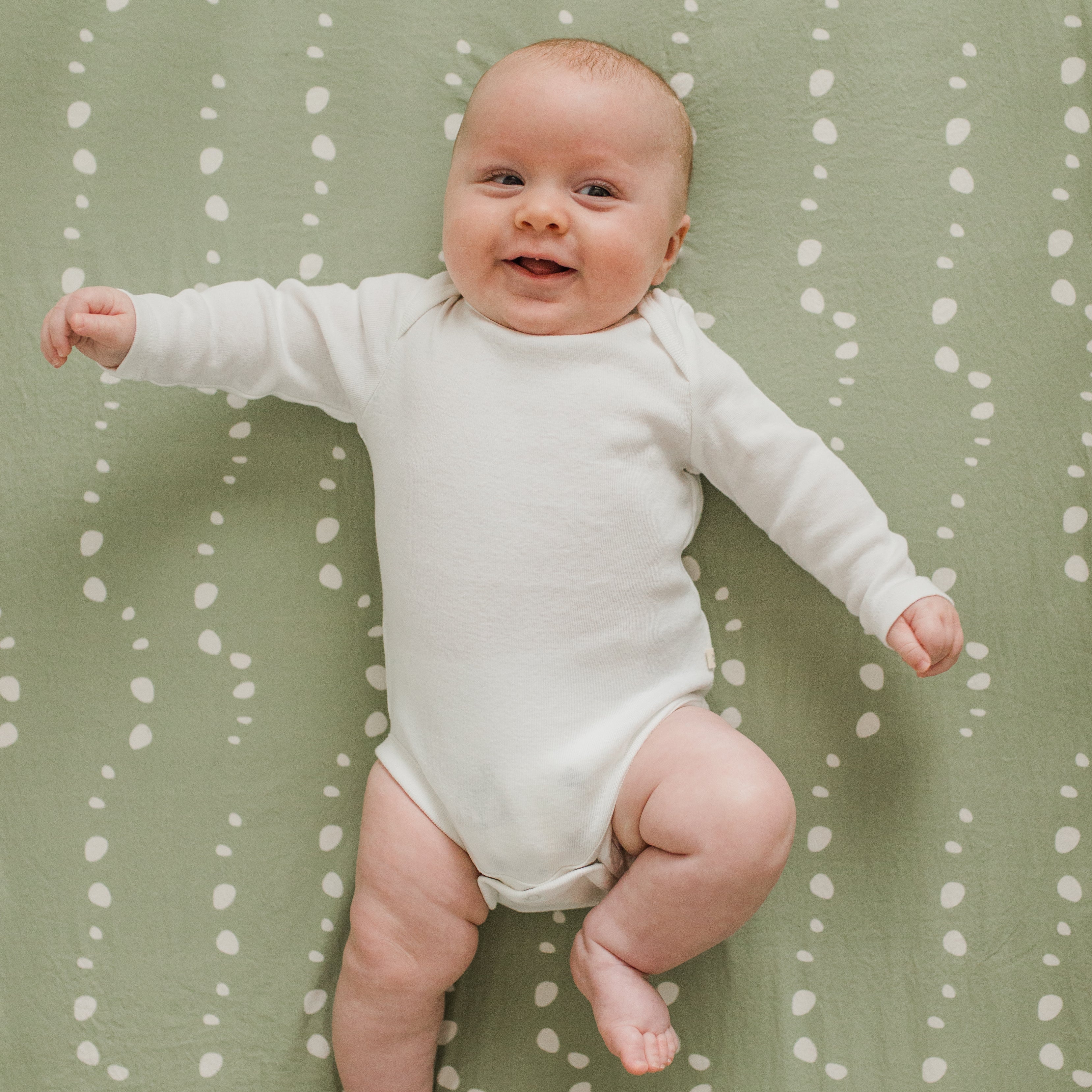 Smiling baby wearing a white onesie, squirming on top of an Oolie organic cotton crib sheet in soft green with a white stones pattern.