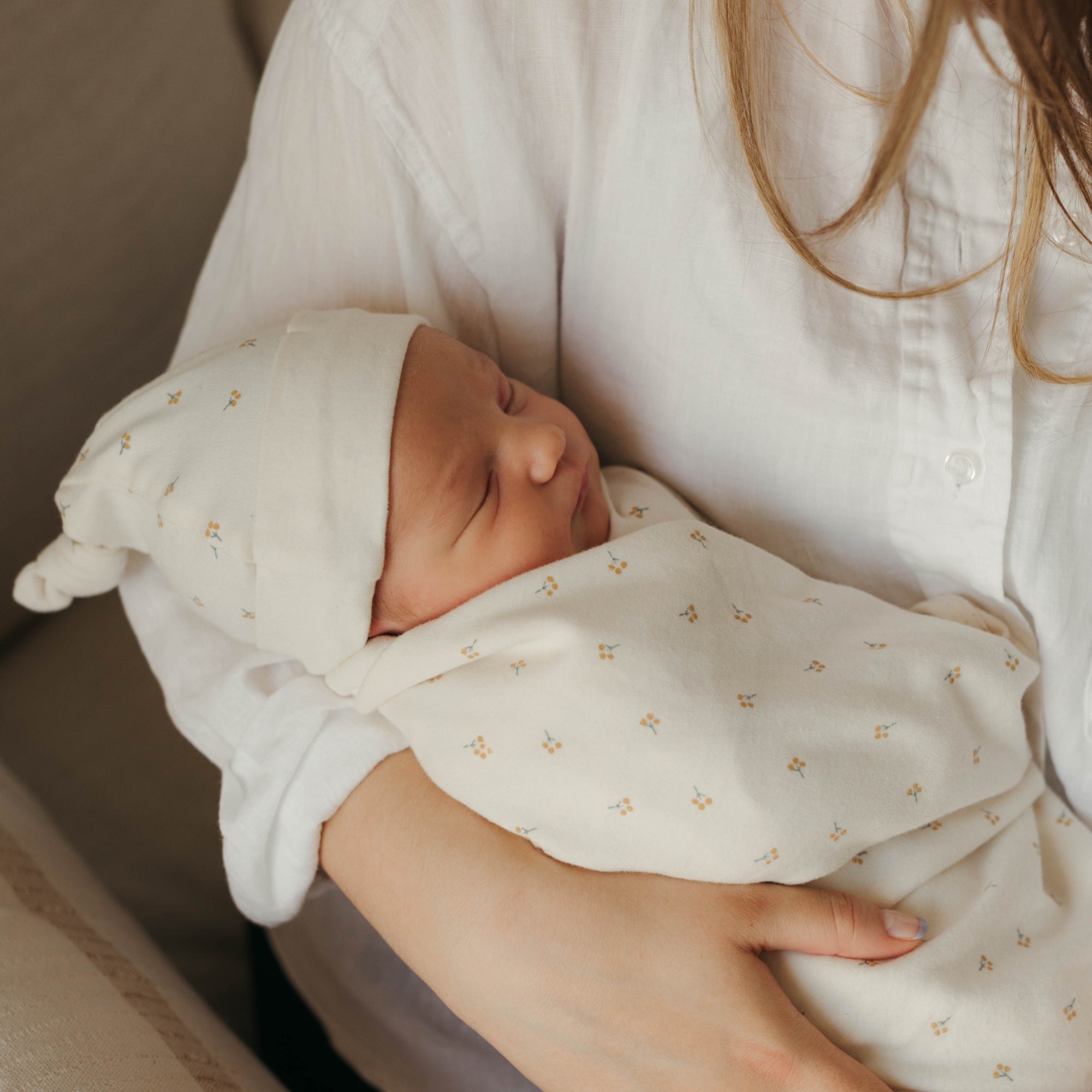 A mom snuggling a newborn baby in her arm. The baby is wearing an organic cotton baby hat from Oolie, and is swaddled in an organic cotton, natural color blanket. Both the blanket and the hat have a repeating printed pattern of a small sprig with golden berries.