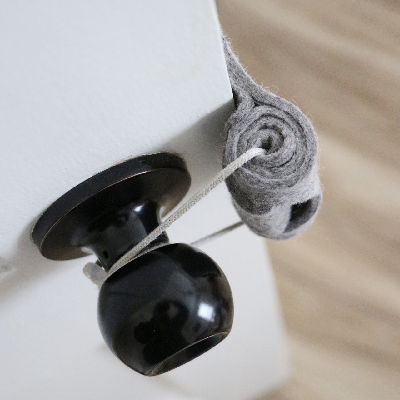 The Oolie Shoosh door silencer and safety bumper shown installed on a door in silencing mode. A gray cord is snugly looped around the door handle, holding the felt bumper in place.