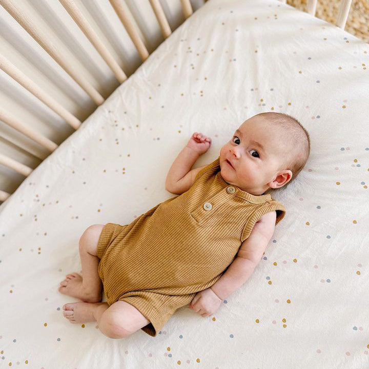 A curious baby looking up at the viewer from his crib, wearing a yellow, corduroy onesie on top of an Oolie organic cotton crib sheet with the cream confetti print, a natural color with small, irregular spots of color.