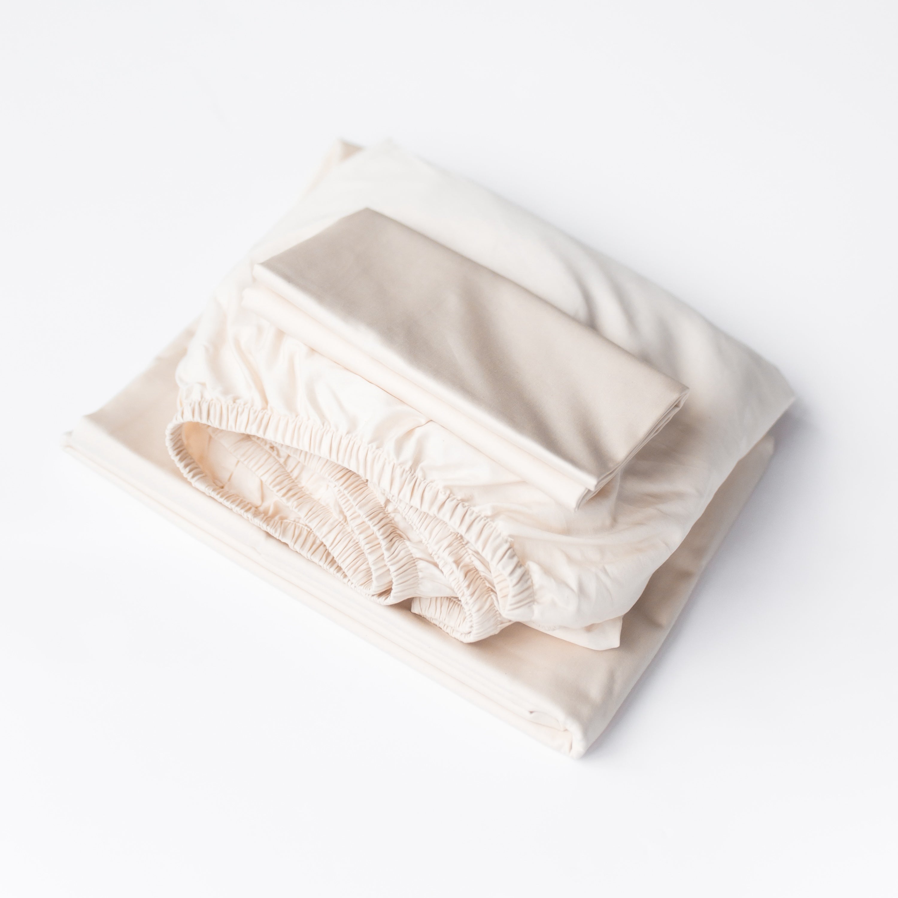 Oolie organic cotton pillowcases, flat sheet, and fitted sheet in natural, unbleached cotton, folded flat and stacked on a white background.