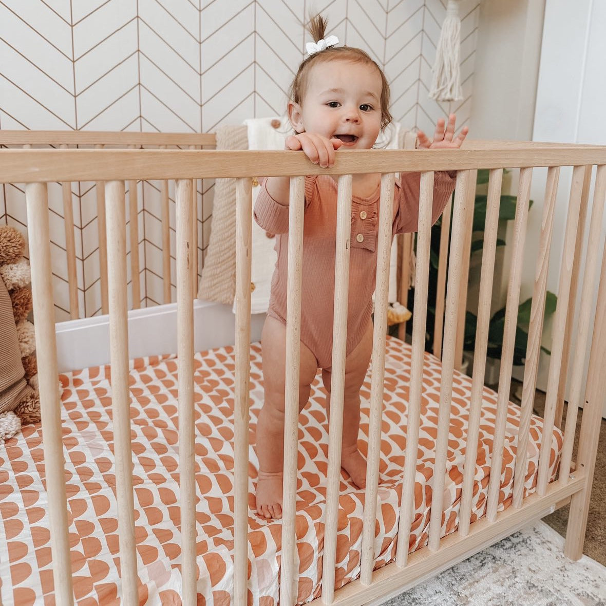 A smiling baby, standing up in her crib, wearing a dusty pink onesie, on top of an Oolie organic cotton crib sheet with the red hills print, a natural color with a series of repeating red semicircles.