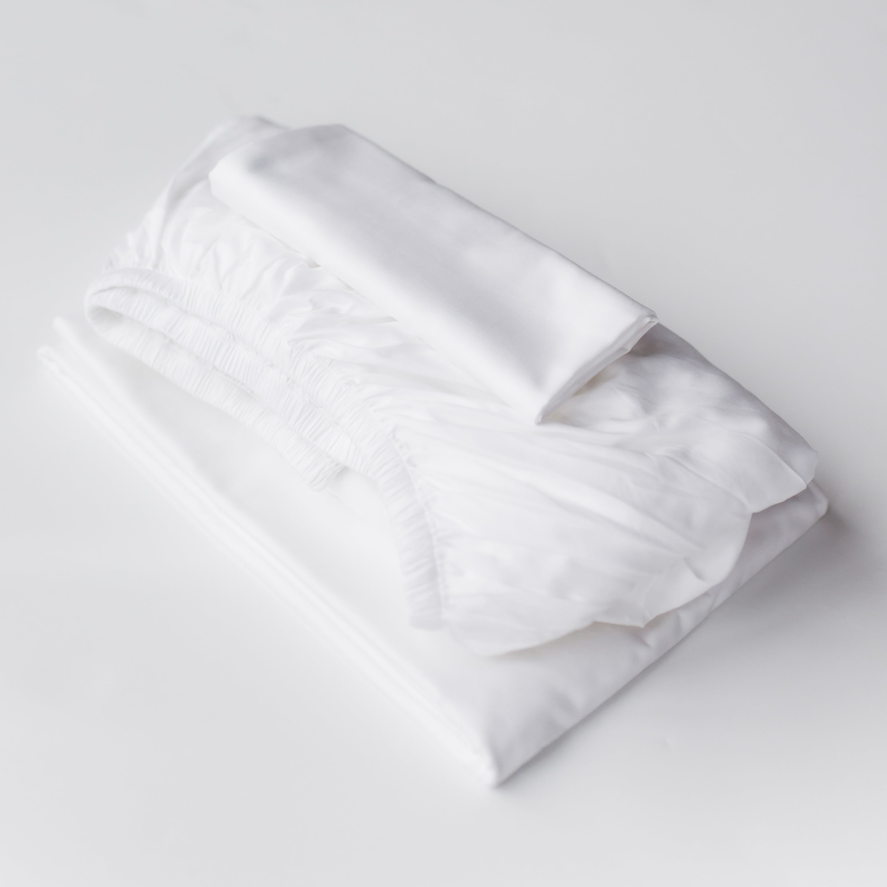 Oolie organic cotton pillowcases, flat sheet, and fitted sheet in white cotton, folded flat and stacked on a white background.