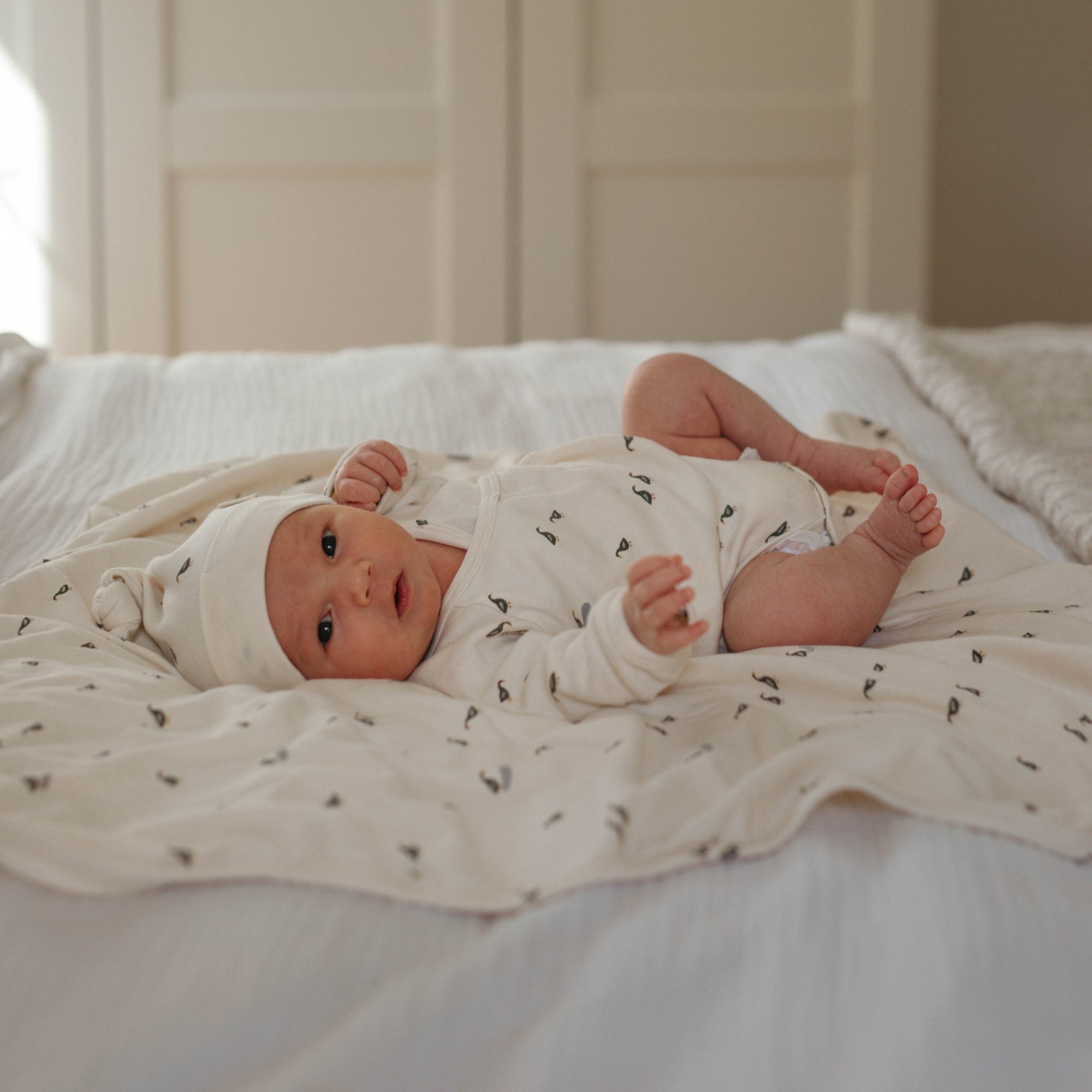 A newborn lying down on an organic cotton baby blanket from Oolie, and looking at the camera. The baby is wearing an organic cotton hat and bodysuit. The hat, bodysuit, and blanket all have a repeating printed pattern of small ducks.