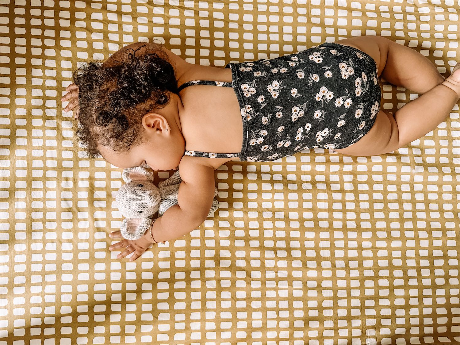 A sleeping baby snuggling a plush elephant toy, sleeping on an organic cotton Oolie crib sheet with a yellow gridded pattern