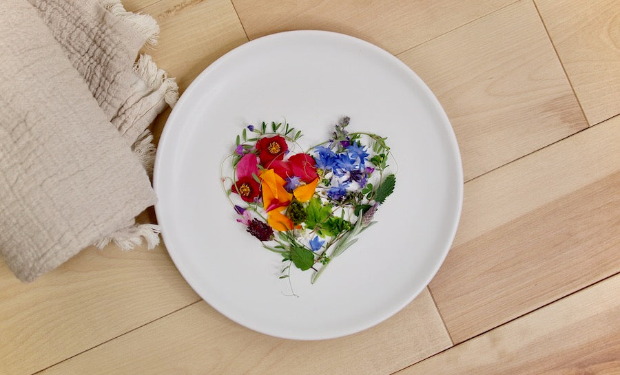 Flower petals arranged in the shape of a heart on a white plate, resting on a natural wood surface, with a natural blanket nearby