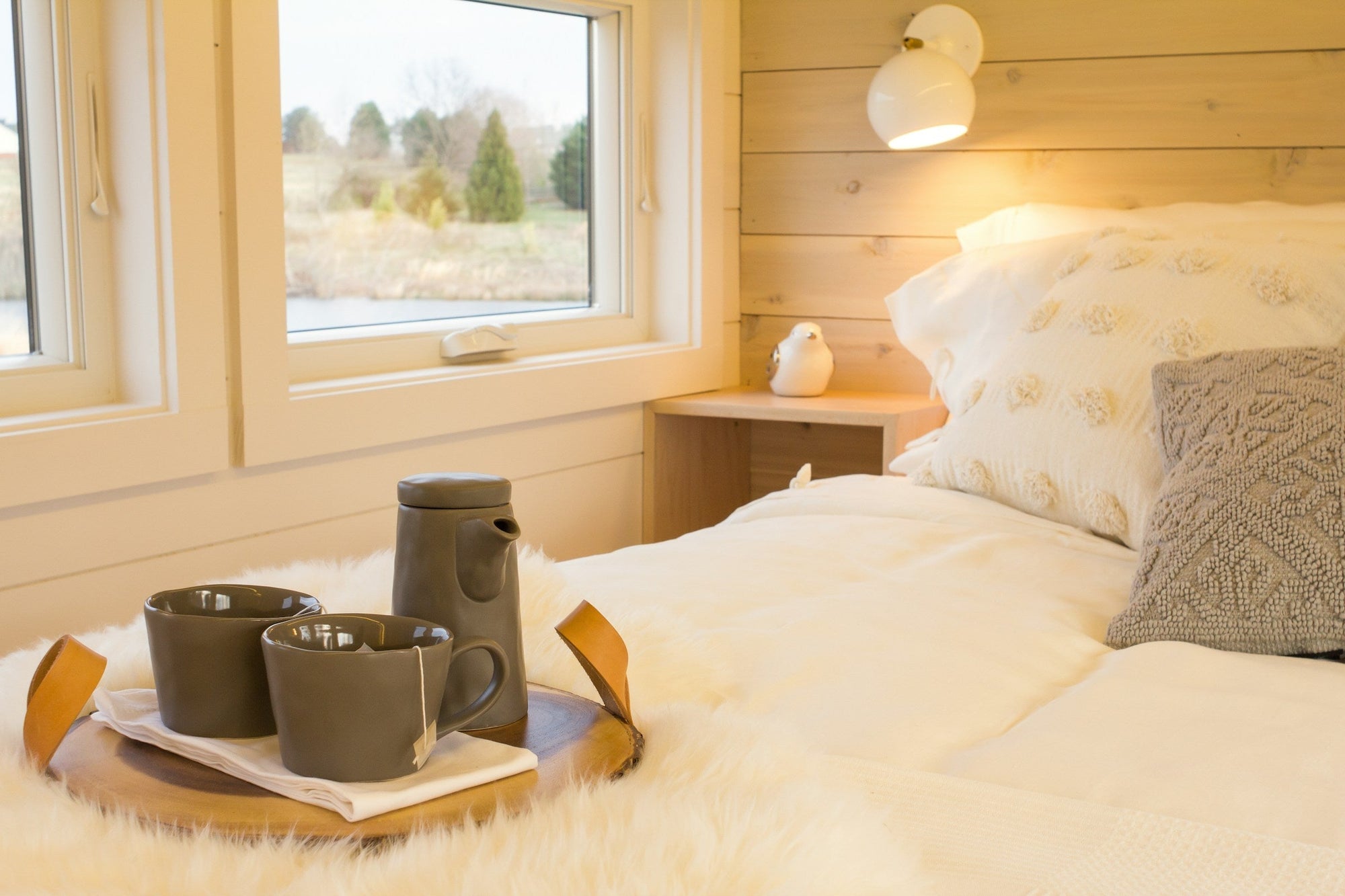 Warmly made bed with soft blankets and pillows in a modern wooden cabin, with a tray with hot tea set on the bed, and grassy woods visible out the window in the background.