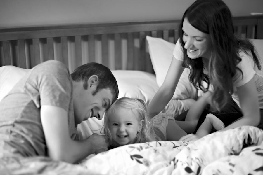 A mom and dad with their young daughter, smiling and laughing together in bed.