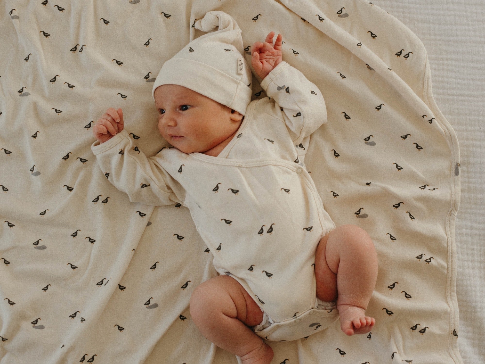 Newborn baby wearing Oolie organic hat and bodysuit, on top of an Oolie organic baby blanket, with the runner duck print.