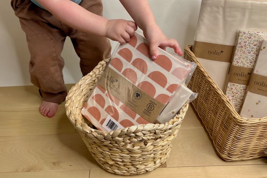 A toddler's hands packing Oolie organic cotton crib sheets into a natural, woven basket.