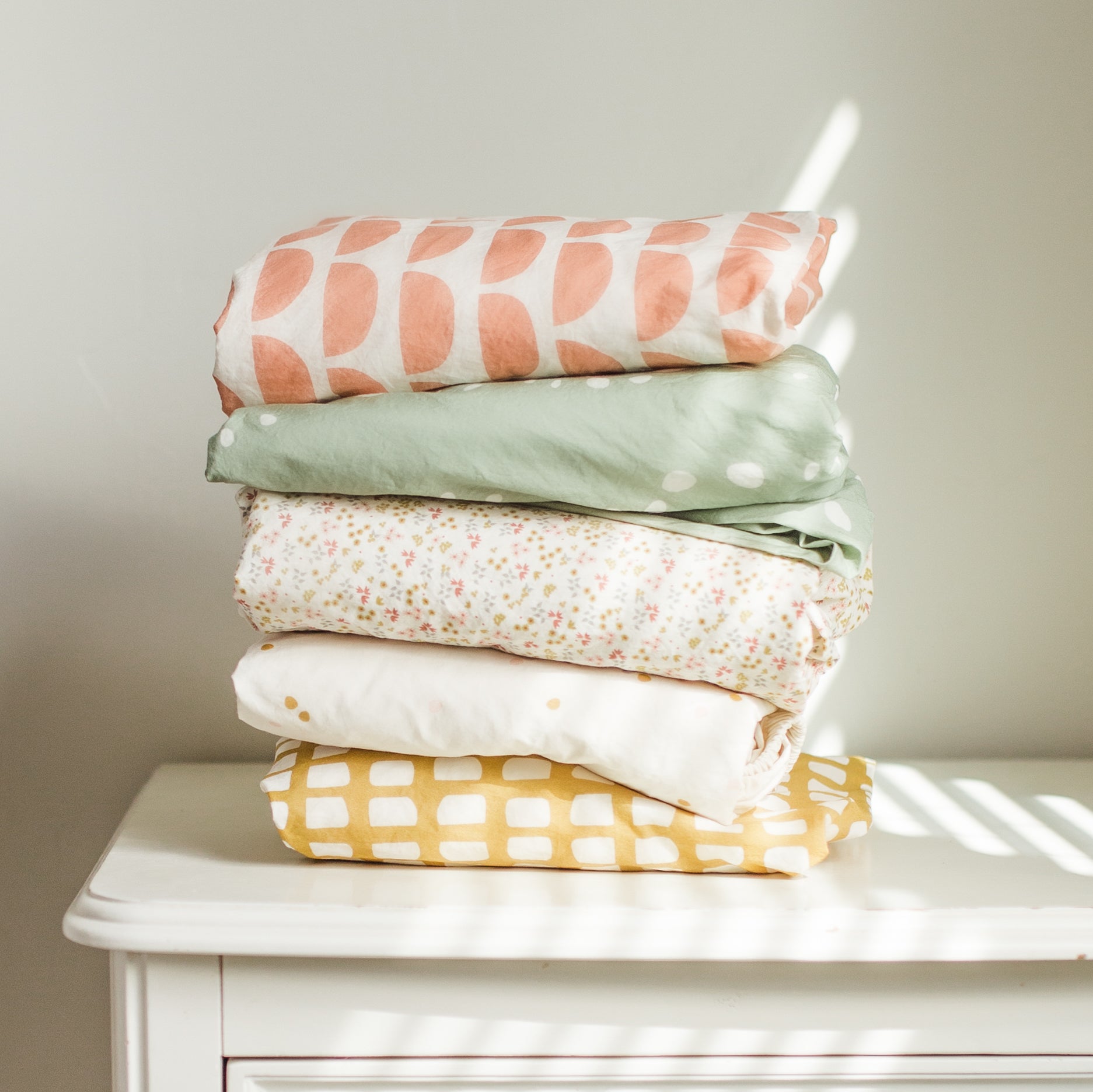 Oolie organic cotton crib sheets in five different graphic prints, gently folded in a stack.
