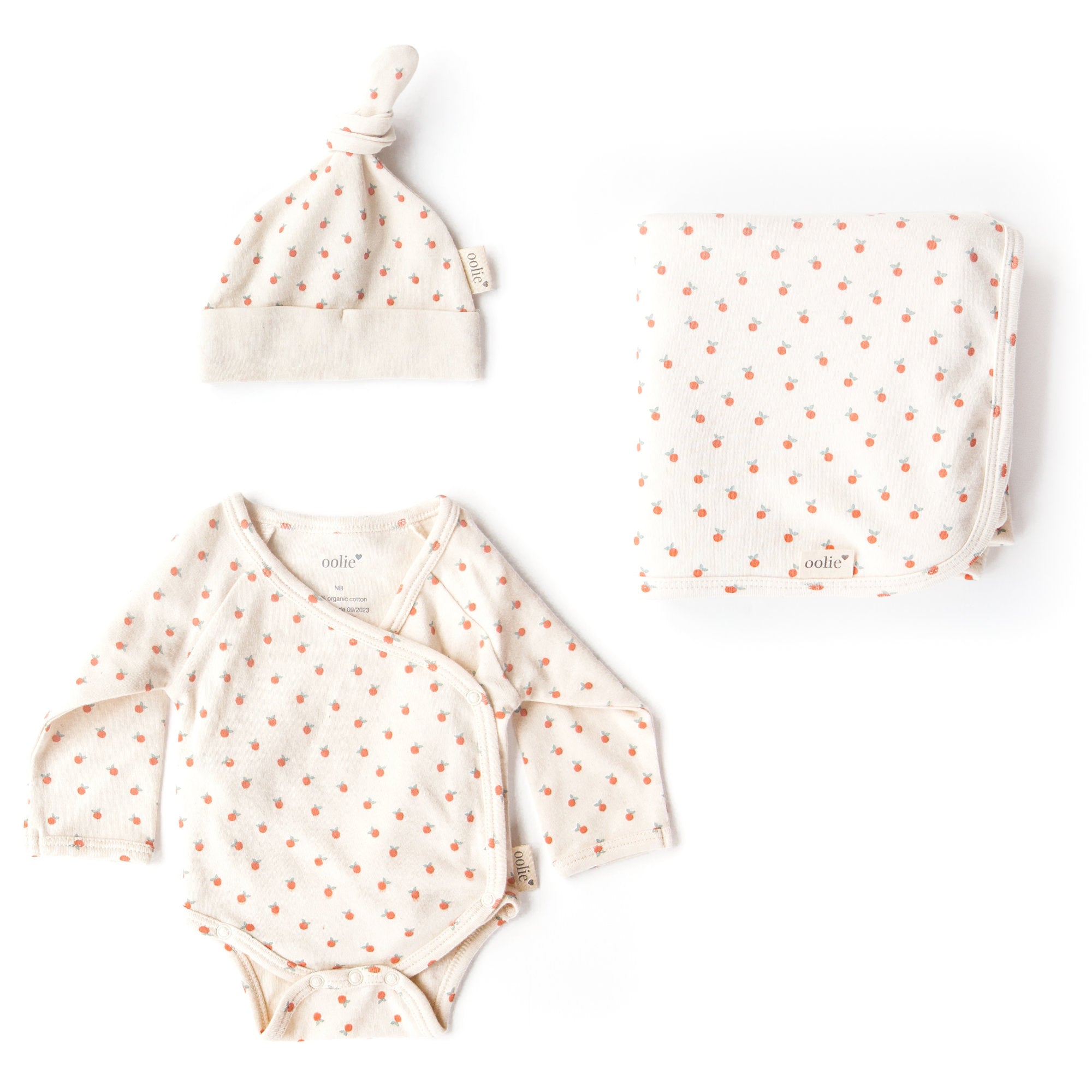 Oolie organic baby hat, baby bodysuit, and baby blanket, laid flat on a white background, each with a little peach print, a natural color with tiny, irregular peaches in a repeating pattern.