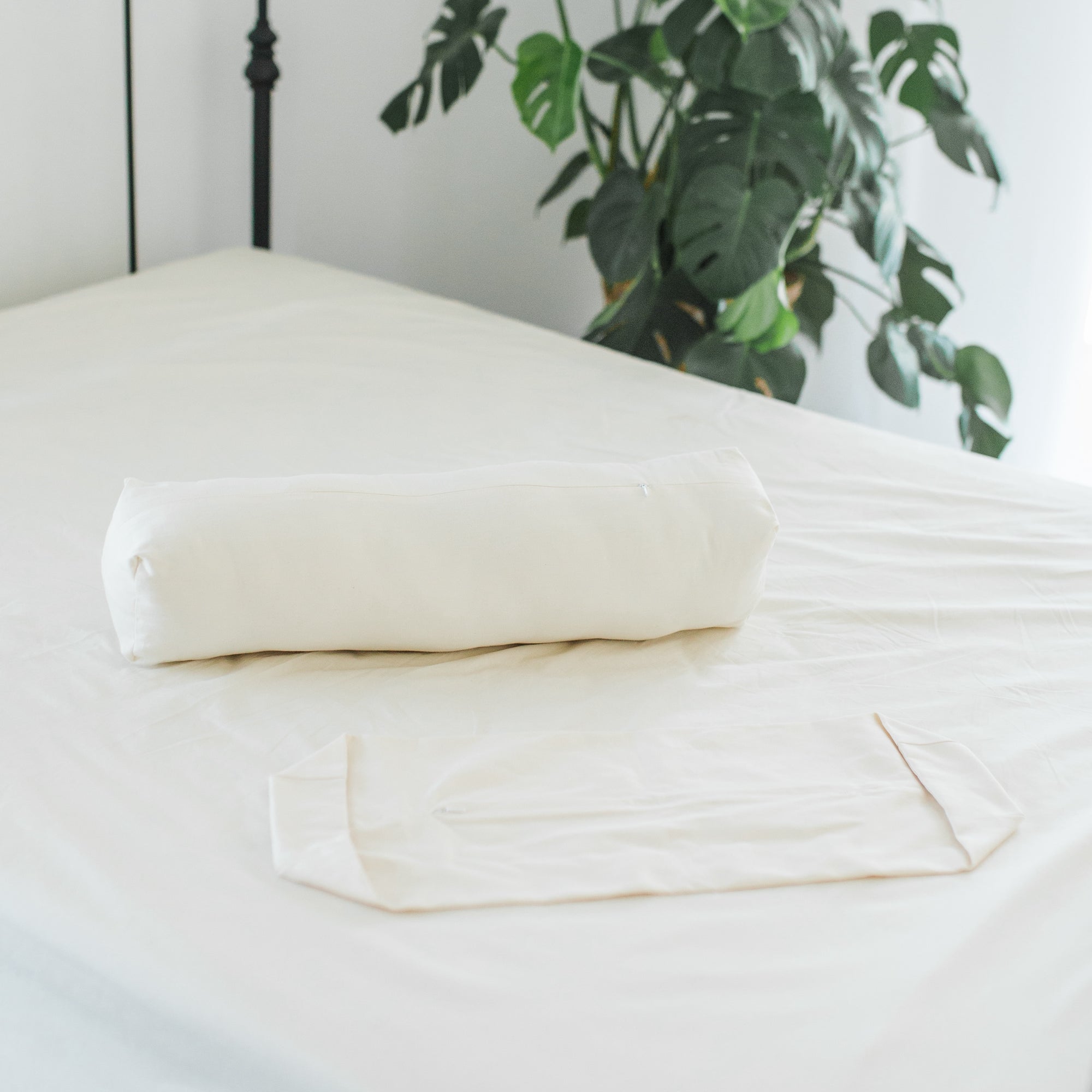 Two short-length Oolie organic cotton bolster pillows, one filled and one unfilled, arranged on a bed.