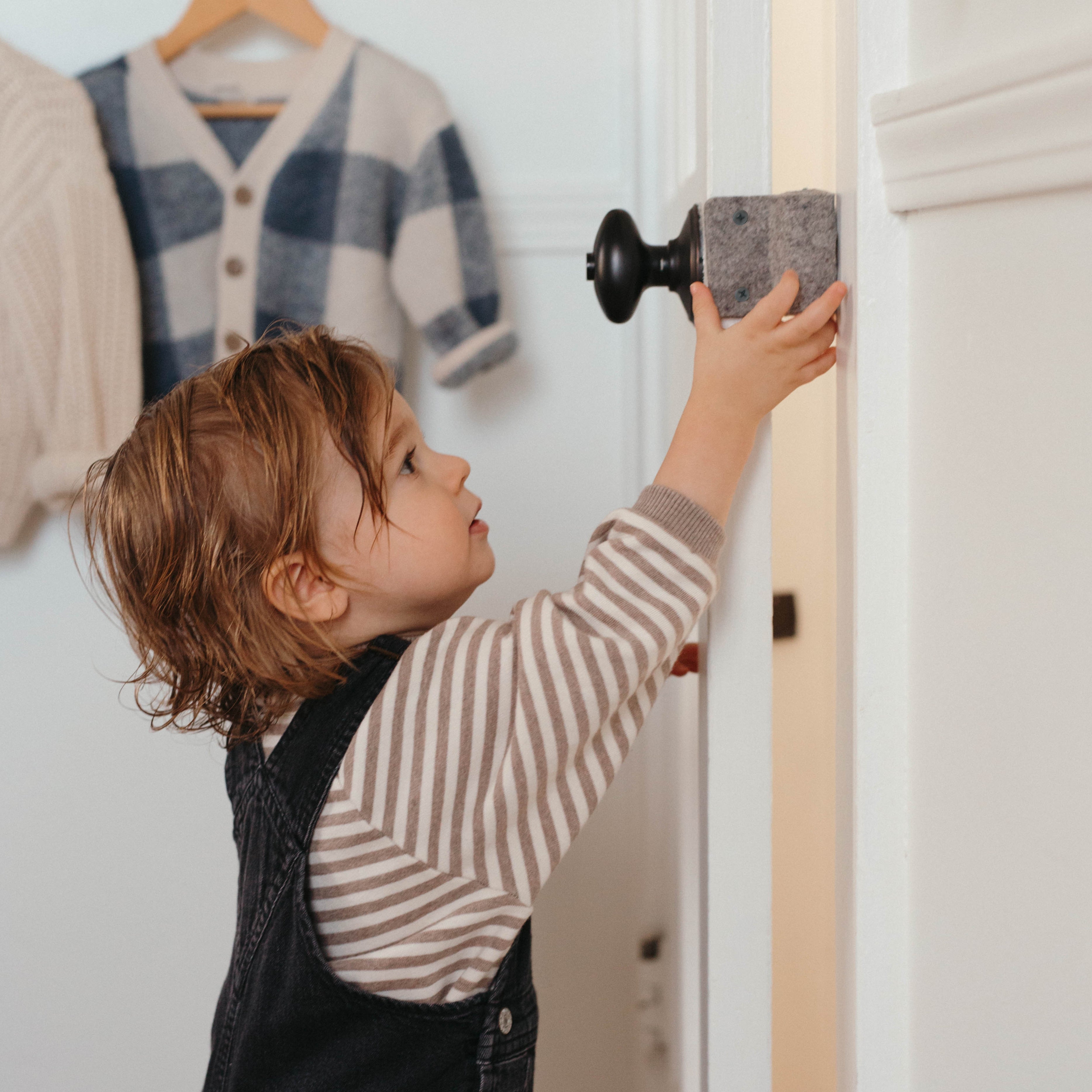 A toddler reaching up to grasp the Oolie Shoosh door silencer and safety bumper, shown installed in Bumper Mode, which prevents the door from closing fully, thereby protecting children's fingers from getting pinched in the door.