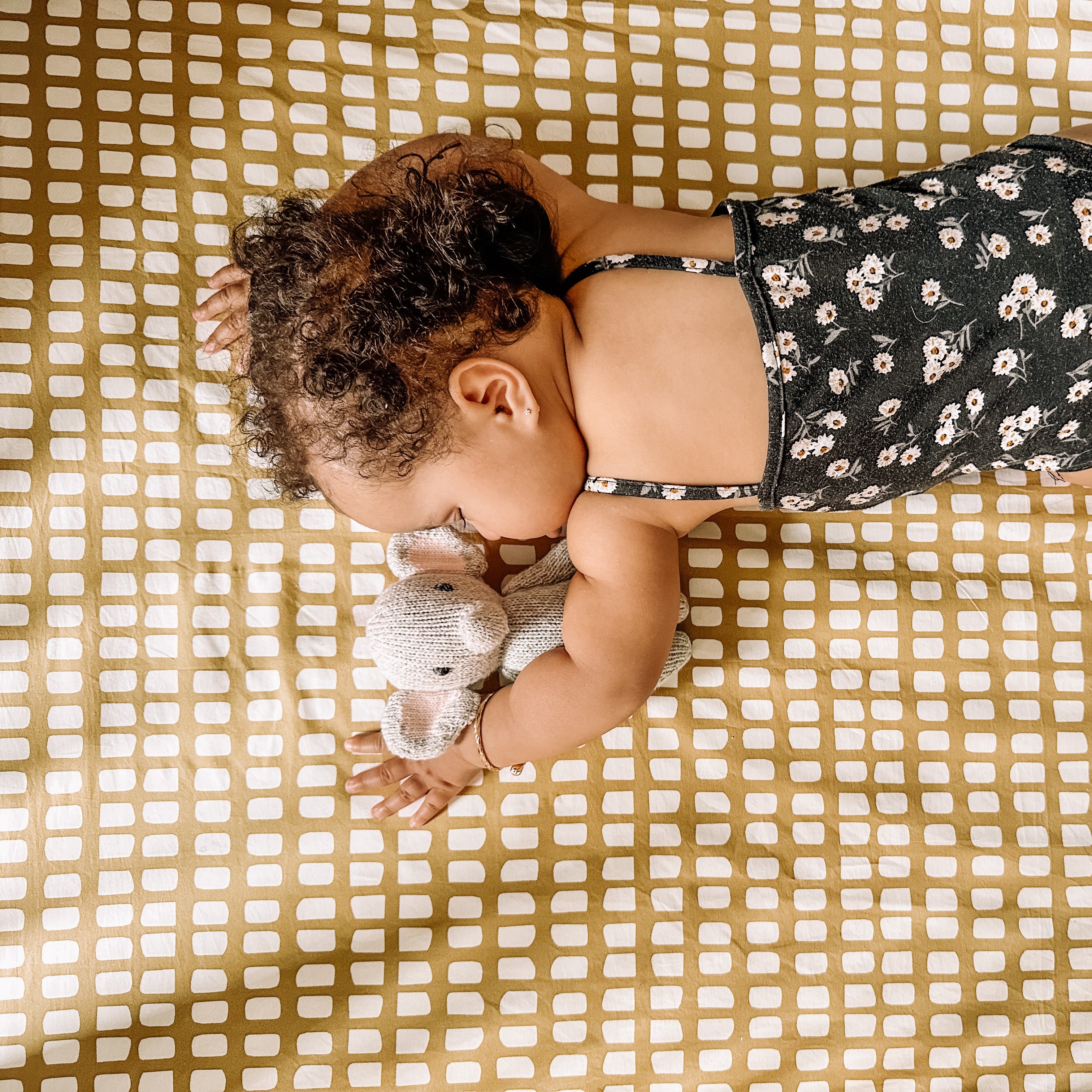 A sleeping baby wearing a black floral onesie, snuggling an elephant toy on top of an Oolie organic cotton crib sheet in deep gold with irregular panels in white.