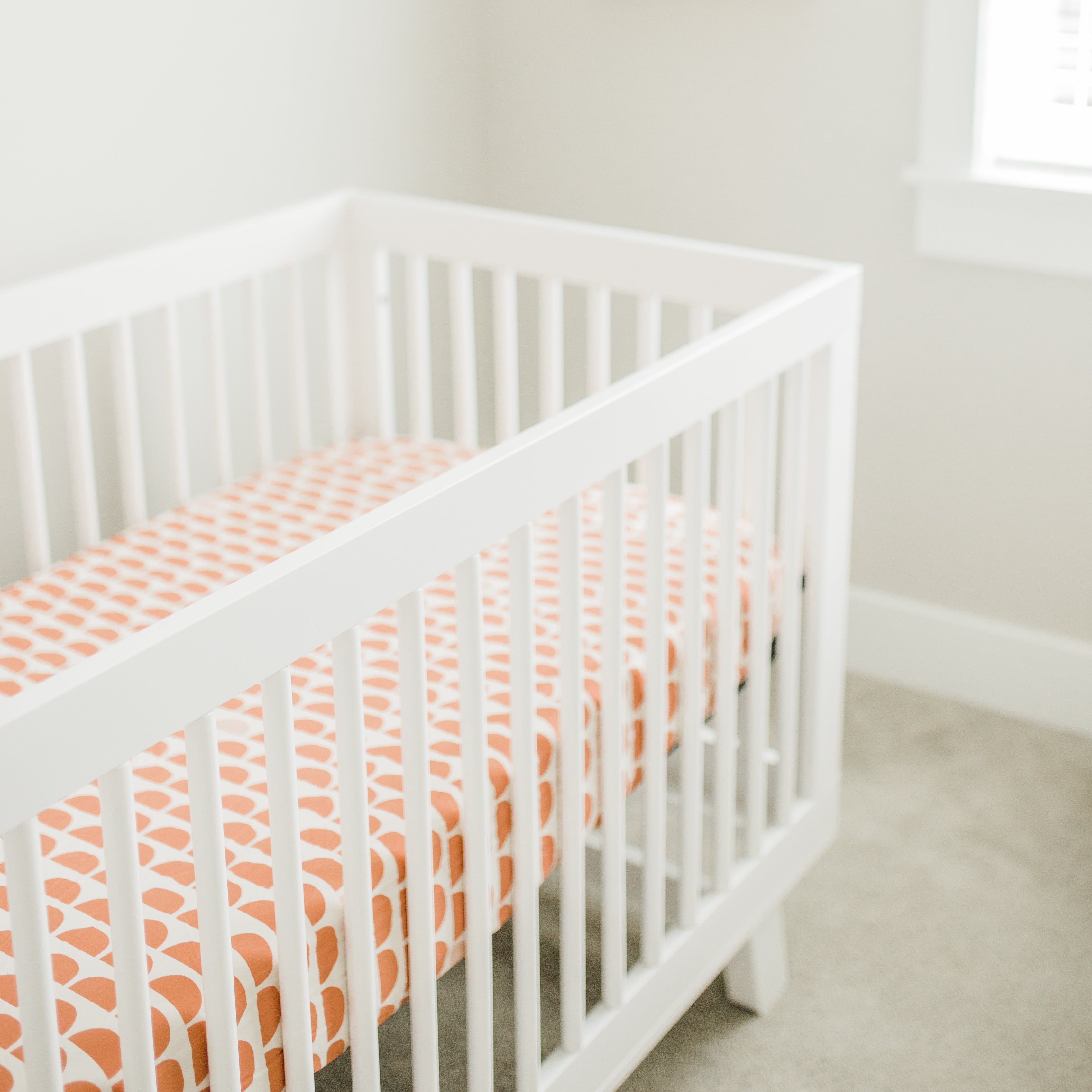 An Oolie organic cotton crib sheet with red graphic print installed on a mattress in a white crib in a baby's room.