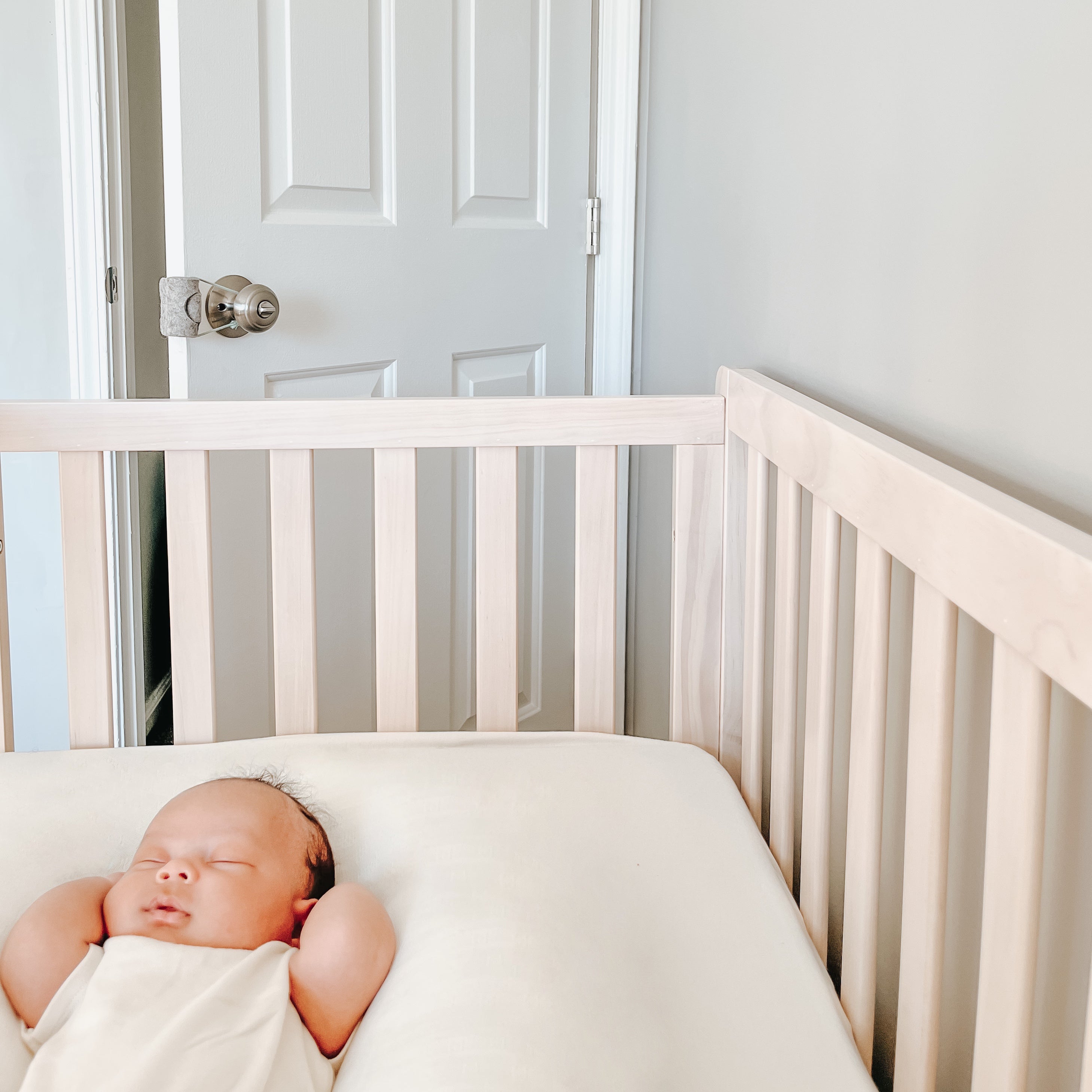 A baby sleeping in a crib, with the door ajar in the background. On the door is installed an Oolie Shoosh door silencer and safety bumper shown, with a light blue cord snugly looped around the door handle, holding the felt bumper in place.
