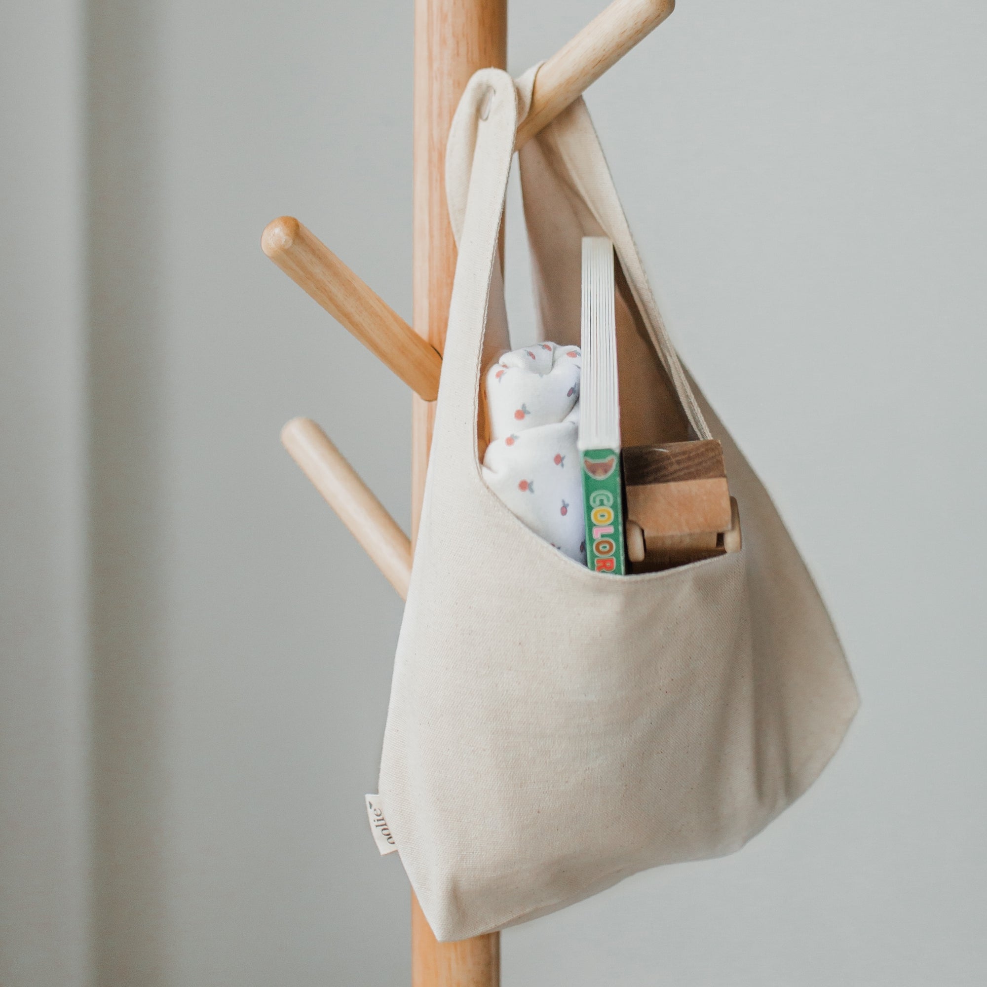 Oolie's convertible, organic bag made of natural color canvas, with handles snapped together, hanging on a wooden coat rack, containing a baby blanket, a small book, and a wooden toy.