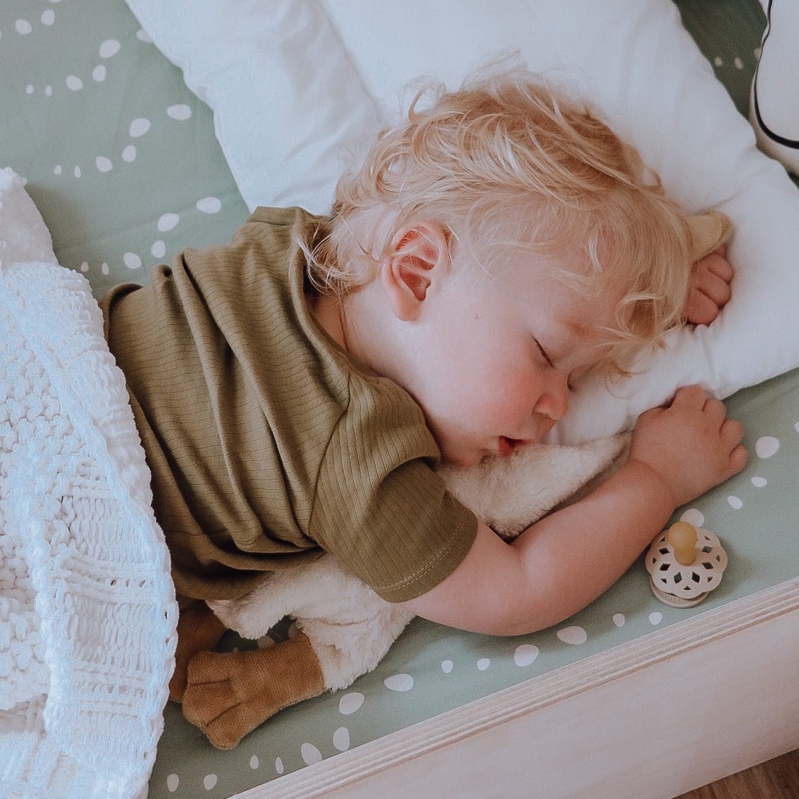 A sleeping baby wearing a yellow onesie, snuggling a duck toy on top of an Oolie organic cotton crib sheet with the sage stones print, a soft green color with white bubbles or irregular stone shapes.