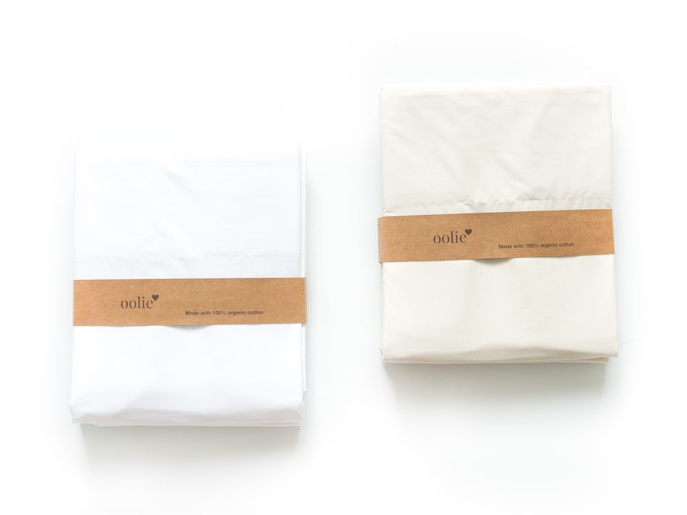Two Oolie organic cotton sheet sets (one white and one in undyed, unbleached natural color cotton), folded, and shown with a kraft paper belly band with the Oolie logo